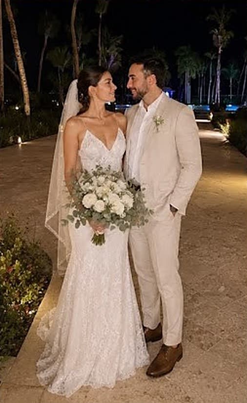 Fernanda con vestido de novia, velo y ramo junto a Diego de traje claro, de noche entre palmeras